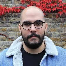 A man with a bald head, beard, and round glasses is wearing a light blue jacket with a fleece collar. He stands in front of a stone wall with red ivy growing along the top, appearing ready to discuss Support Resolution via Intercom.