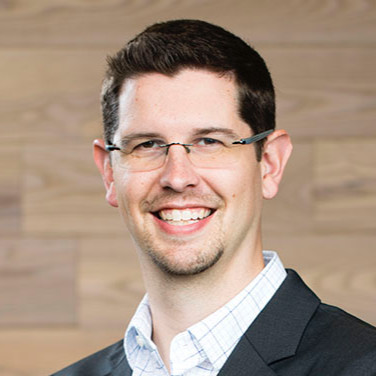 Jon Wishart, a man with short dark hair, glasses, and a trimmed goatee, wearing a dark blazer and light checkered shirt, smiles in front of a light wooden wall background. He’s known for his insights on B2B Communities.