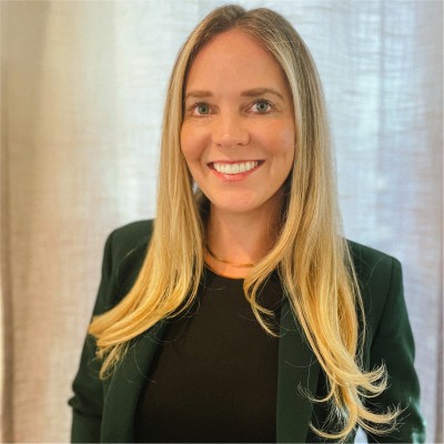 A woman with long blonde hair, wearing a dark green blazer over a black top, is smiling at the camera. She stands in front of light-colored curtains, embodying the spirit of category creation embraced by innovators at Gainsight.