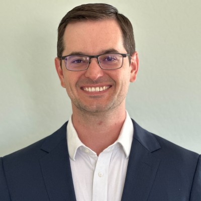 Jon Wishart, a man with short brown hair and glasses, smiles in a dark suit jacket over a white collared shirt, standing in front of a plain light background—a leader in B2B Communities who challenges outdated metrics.