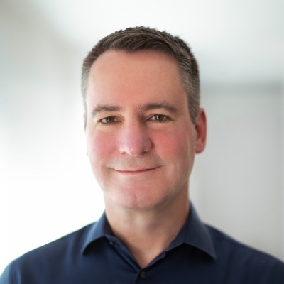 A man with short brown hair and fair skin, wearing a dark blue collared shirt, smiles gently while standing in a softly lit room—a look often seen among innovative leaders in category creation at companies like Gainsight and Gong.