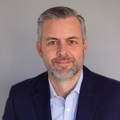 A middle-aged man with short gray hair and a beard, wearing a dark suit jacket over a light blue checkered shirt, poses in front of a plain gray background—Jared Collins of $100B Company leading the way in Consumption-Based CS.