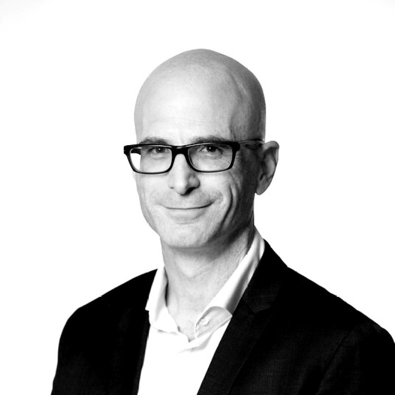 A bald man wearing dark-rimmed glasses, a suit jacket, and a collared shirt—reflecting the professionalism of the Obrela CS team—smiles slightly while facing the camera against a plain white background. The image is in black and white.