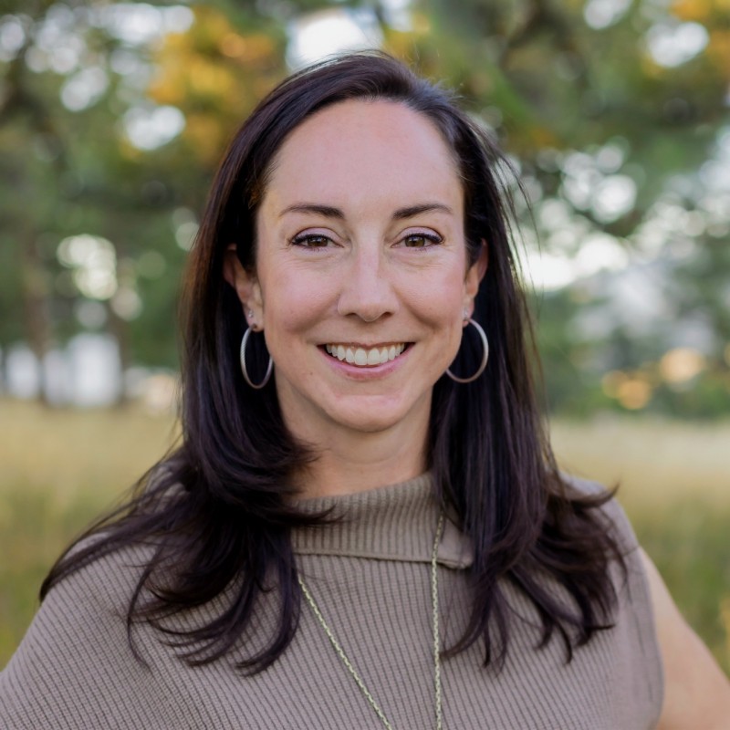 Christine Lavery, with long dark hair, smiles outdoors in a taupe top and hoop earrings. She manages some of the Biggest Accounts at Digital Motions, with blurred trees and greenery in the background.
