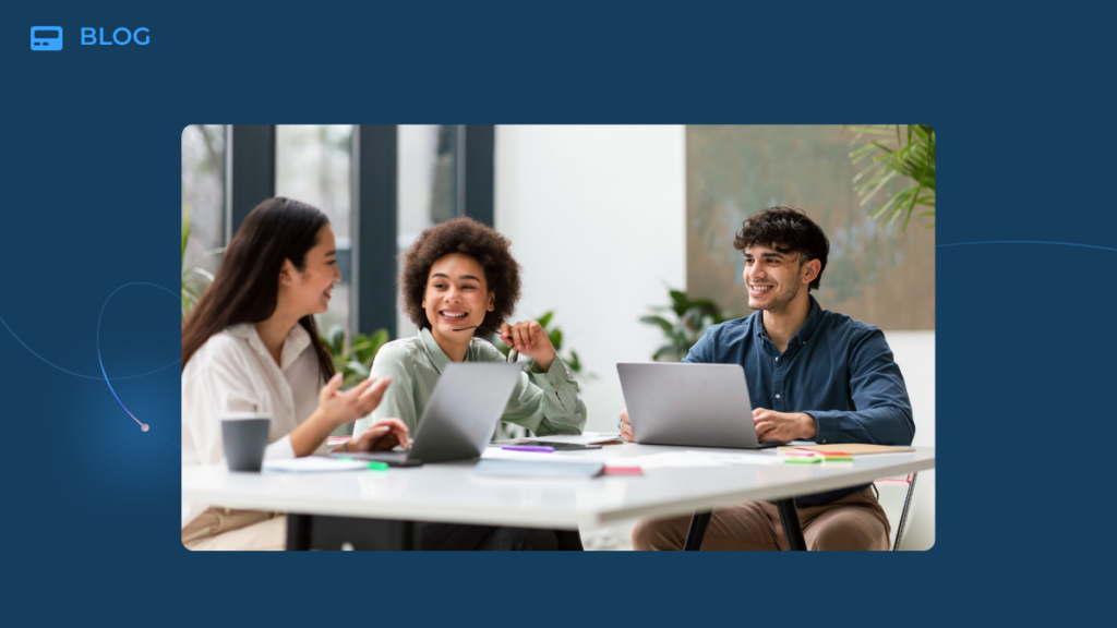 Three people sit at a table with laptops, smiling and talking together in a bright, modern office setting, discussing metrics and building a strong customer community amid plants and coffee mugs.