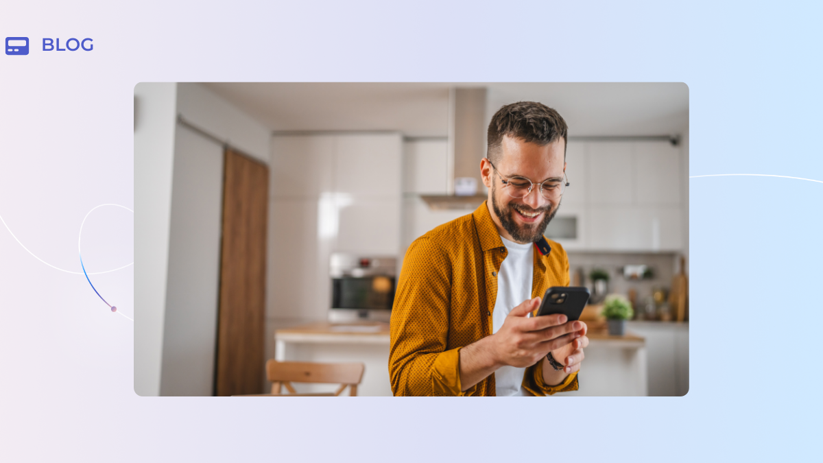 A smiling man with glasses and a beard stands in a modern kitchen, wearing a yellow shirt and looking at his phone—ready to take customer success management to the next level, just like a CSM team at a $100 billion company.
