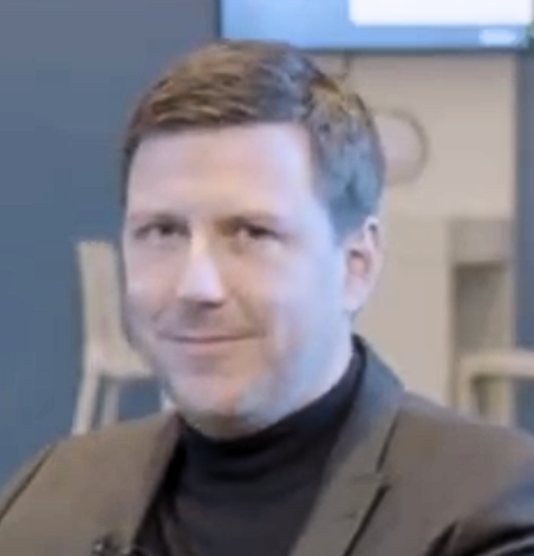 A man with short brown hair, wearing a dark blazer and black turtleneck, sits indoors and smiles slightly at the camera. The background is blurred with blue walls and a television screen displaying SAP Customer Success Strategy.