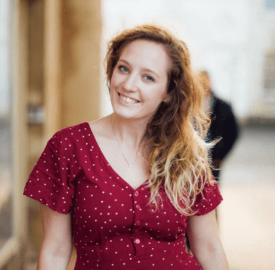 A woman with wavy blonde hair, wearing a red dress with white polka dots, stands outdoors smiling at the camera. The background is softly blurred, with someone behind her discussing AI and support tickets on Slido.