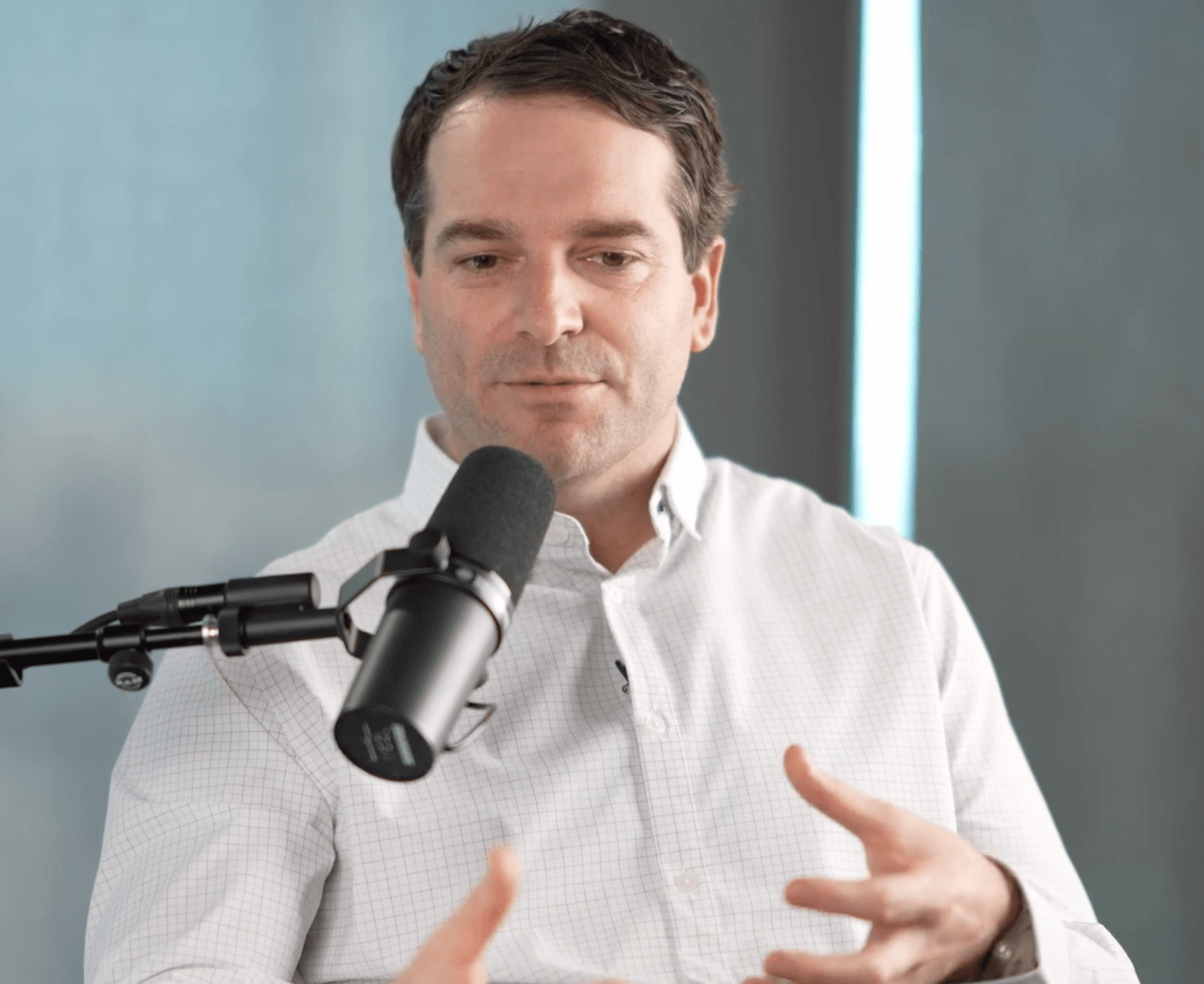 A man in a white button-up shirt speaks into a microphone during a podcast or interview, gesturing with his hands as he discusses customer success. The background is blurred and softly lit.