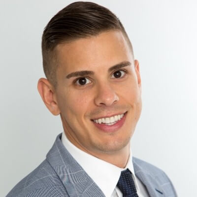 Smiling man with short dark hair, dressed in a light blue suit jacket, white shirt, and dark tie, posing confidently in front of a plain light background—perfect for conveying sales professionalism or showcasing CCO ownership.