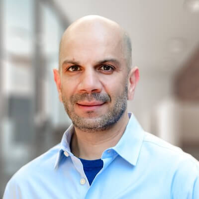 A bald man with light skin, a trimmed beard, and a blue collared shirt smiles softly while standing indoors in a brightly lit, modern setting, reflecting the confident demeanor of a seasoned GTM or CRO leader at Replit.