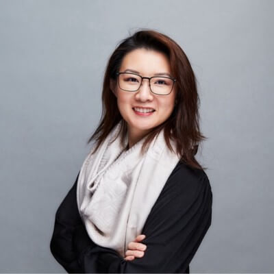 A woman with shoulder-length brown hair and glasses, wearing a black top and a light-colored scarf, stands smiling with her arms crossed against a plain gray background, embodying the confidence of a Gainsight post-sales playbook expert.