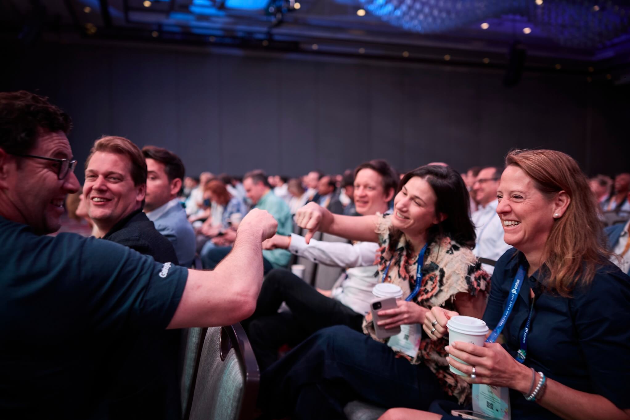 A group of people sitting in an audience laugh and smile while two of them fist bump. Several hold coffee cups, enjoying the lively atmosphere at Pulse 2026 as they await the next speakers on the agenda.