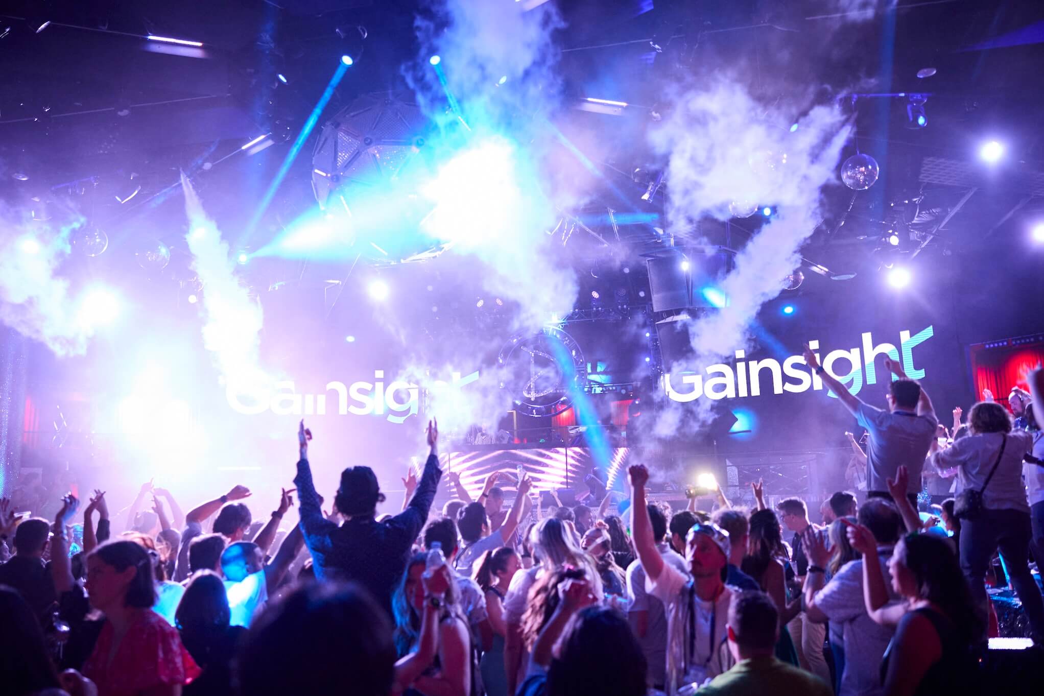 A large crowd dances and cheers in a vibrant nightclub with bright lights, smoke effects, and "Gainsight Pulse 2026" projected in the background. The atmosphere is energetic and festive as speakers engage the audience.