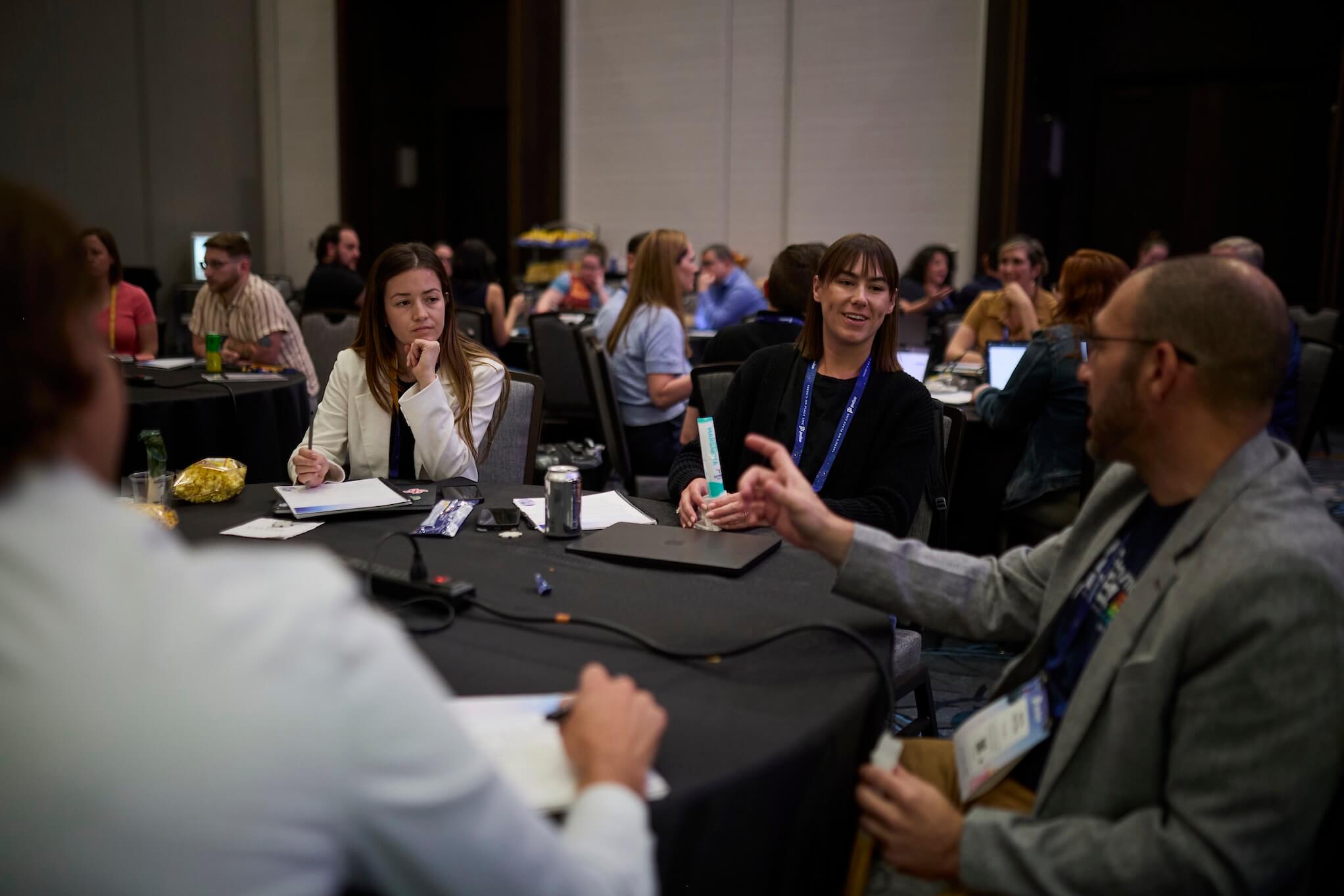 People seated around round tables in a conference room discuss the Pulse 2026 agenda. Some take notes, while others listen and speak. The atmosphere is collaborative and professional as speakers engage with participants.
