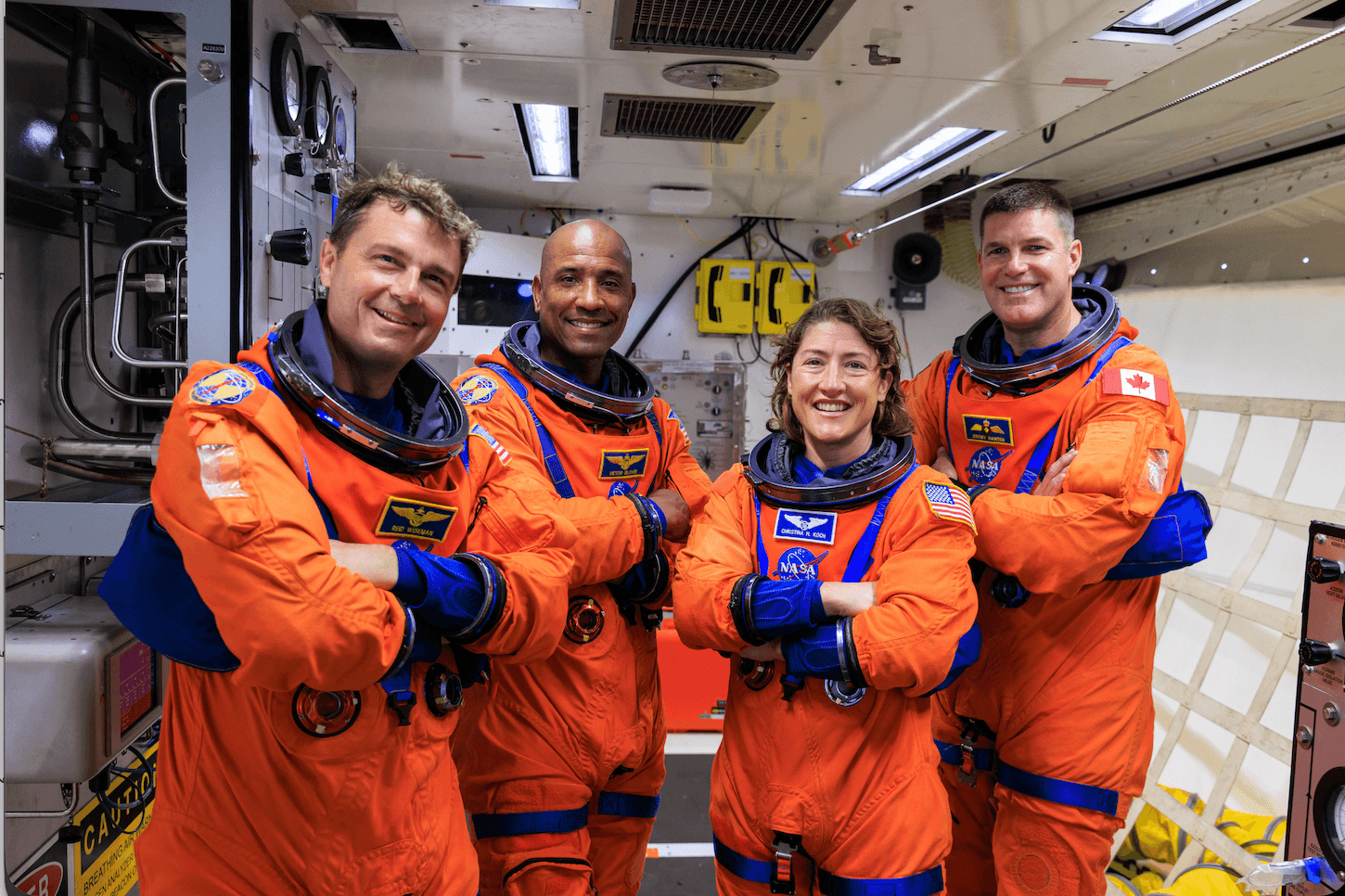 Four astronauts in orange NASA spacesuits stand side by side inside a spacecraft, smiling with arms crossed. Behind them, equipment and control panels hint at the mission's Pulse 2026 agenda.