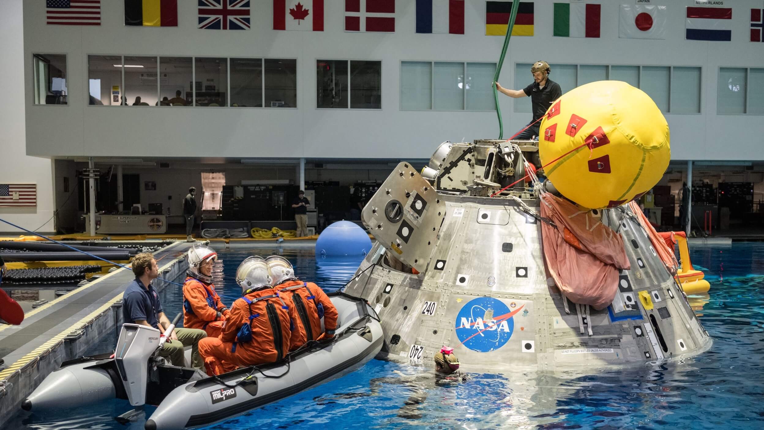Astronauts in orange suits sit in a raft next to a floating NASA capsule in a pool, with a person on top of the capsule holding a yellow buoy. International flags hang above large windows—hinting at an agenda as global as Pulse 2026.