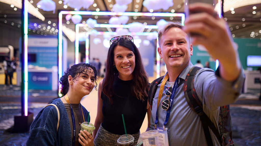 Three people smiling and taking a selfie while attending a vibrant indoor event, holding drinks. Colorful lights and decorative hanging elements set a lively scene—like the energy found at top 2026 Customer Experience Conferences.
