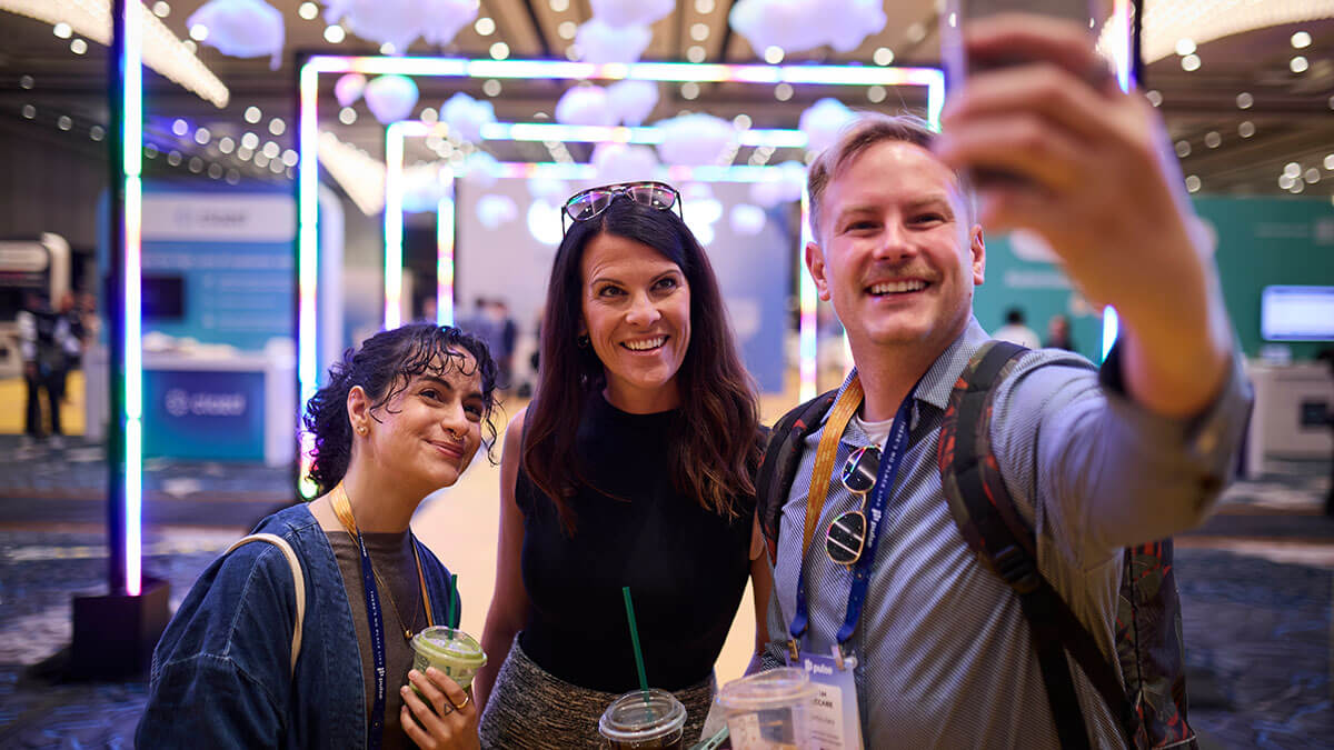 Three people smiling and taking a selfie while attending a vibrant indoor event, holding drinks. Colorful lights and decorative hanging elements set a lively scene—like the energy found at top 2026 Customer Experience Conferences.