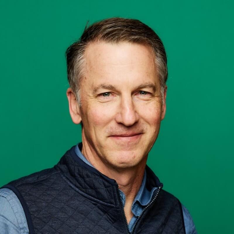 A middle-aged man with short gray hair, Rob Edmondson, smiles softly at the camera. He is wearing a blue collared shirt and a dark quilted vest, standing against a solid green background at Ironclad, where AI predicts churn.