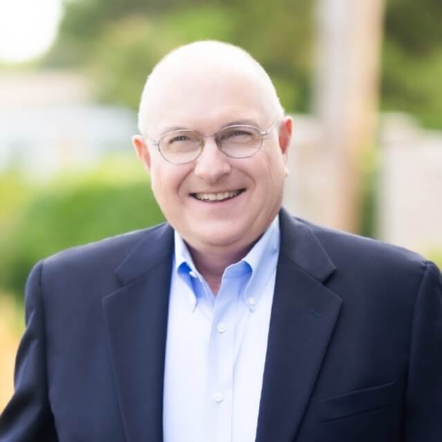 A smiling, bald man with glasses—Fred Reichheld of Bain & Company—wears a navy blazer and light blue shirt as he stands outdoors with a blurred green background, reflecting the innovative spirit behind NPS.