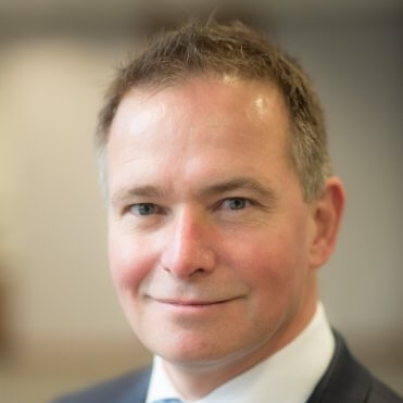 A middle-aged man with short brown hair, light skin, and blue eyes is wearing a dark suit and white shirt, smiling slightly—resembling Jean de Villiers—with a blurred indoor background suggesting an AI-driven workspace at Unit4.