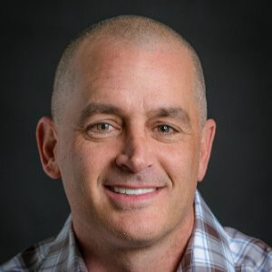 A smiling middle-aged man with a shaved head, wearing a plaid shirt, stands against a dark, plain background—an approachable face often seen leading CS Teams in Commercial Line projects.