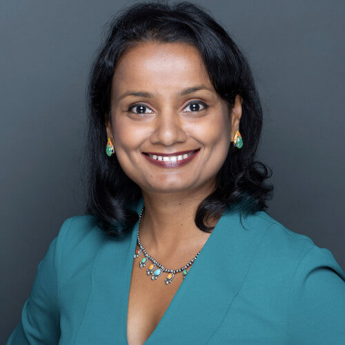 A woman with medium-length black hair, wearing a teal blazer, matching earrings, and a necklace, smiles in front of a plain grey background—articulate and confident, she embodies the spirit of 133 Million Learners.