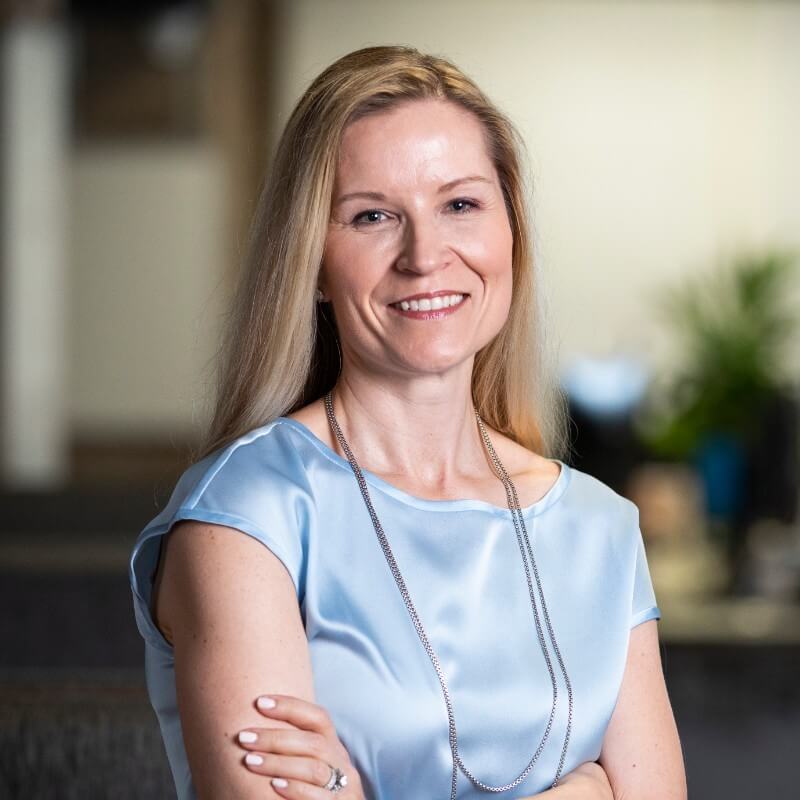 A woman with long blonde hair, wearing a light blue blouse and a long silver necklace, smiles with her arms crossed in an indoor office setting, ready to share insights about LinkSquares product demos.