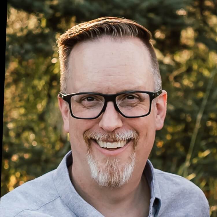 A smiling man with short brown hair, glasses, and a goatee, wearing a light gray shirt, stands outdoors with blurred greenery in the background—Jason Dunn from the AWS Developer Community.