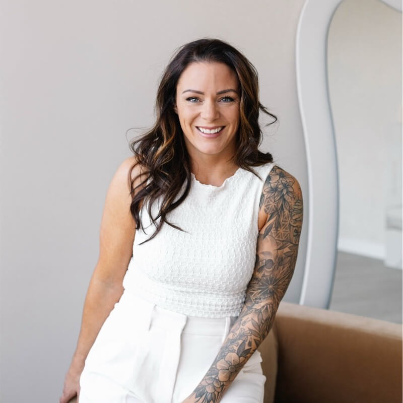 A woman with long brown hair, a large floral tattoo on her arm, and an articulate smile sits on a brown sofa in white attire, with a mirror and light-colored wall behind her.