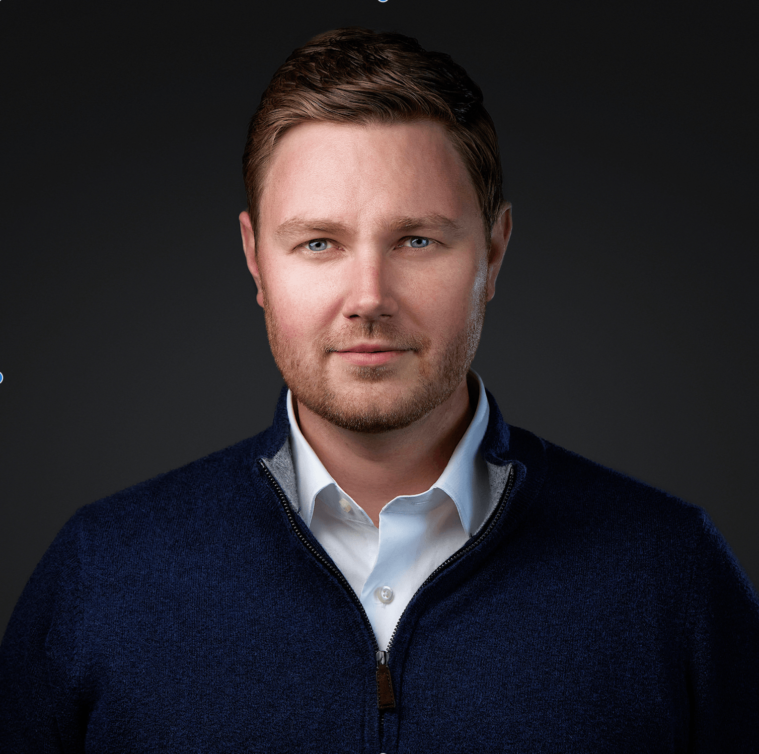 A man with short brown hair and light stubble wears a navy blue zip-up sweater over a white collared shirt. He faces the camera with a neutral expression, embodying the professionalism found in Smart Teams against a dark, plain background.