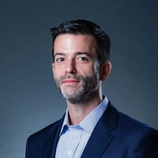 Man with short dark hair and beard wearing a navy suit jacket and light checkered shirt, posing in front of a plain dark background, reflecting a sharp professionalism suited for leading Org Redesign or streamlining Dev Cycles.