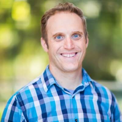 A smiling man, Chris Catania, with short light brown hair wearing a blue plaid shirt stands in front of a blurred green outdoor background, embodying the spirit of community.