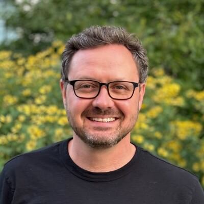 A smiling man with short brown hair, glasses, and a beard is wearing a black t-shirt. He stands outdoors in front of green foliage and yellow flowers, appearing ready to share his latest thoughts on CS Insights.