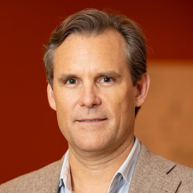 A man with short, light brown hair wearing a tan blazer and light blue shirt stands in front of a blurred, warm brown background, embodying the professional presence of a Chief AI Officer at BCGx.