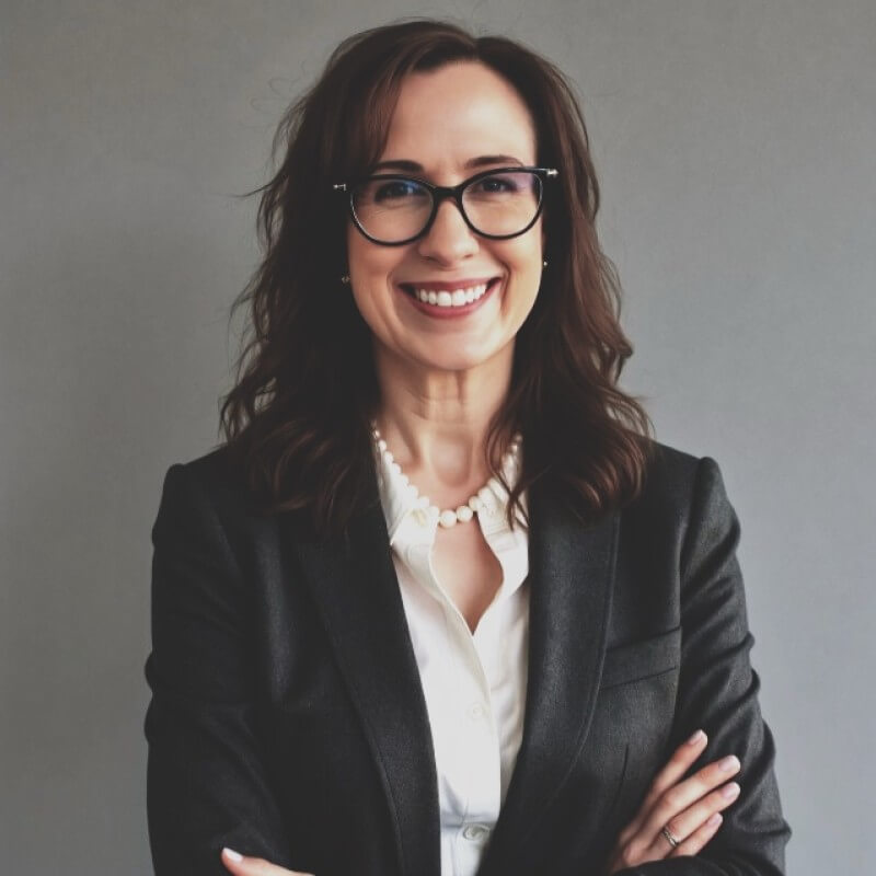 Smiling woman with wavy brown hair wearing glasses, a dark blazer, white blouse, and a pearl necklace stands with arms crossed against a plain gray background—confidently ready to embrace the New Playbook.