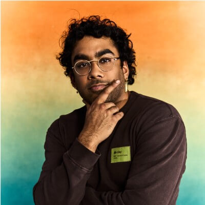A person with glasses and curly hair poses thoughtfully with a hand on their chin, wearing a dark shirt and name tag displaying their job title, set against an orange and teal gradient background.