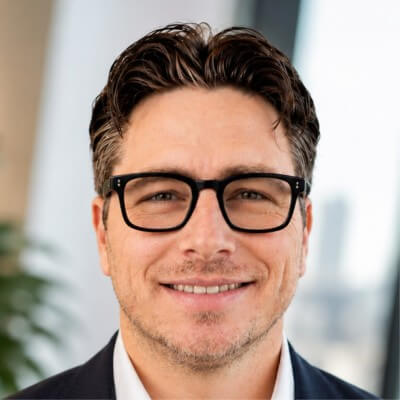 Man with dark, wavy hair and glasses, smiling slightly, wearing a dark suit and white shirt, in a bright office setting with a blurred cityscape in the background—a professional image reminiscent of Ziv Peled, Chief AI Officer at AppsFlyer.