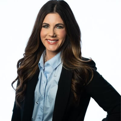 Woman with long brown hair wearing a light blue shirt and black blazer, smiling at the camera against a white background—proving you can stand out in the CS hiring market even without a CS background.