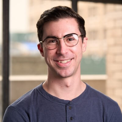 A smiling man with short dark hair and glasses, the CRO responsible for driving revenue at Brex, stands indoors in front of large windows with a blurred cityscape background.