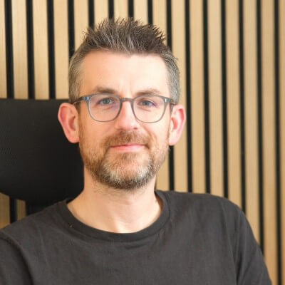 A man with short brown hair, glasses, and a beard, wearing a black t-shirt, sits in front of a wall with vertical wooden slats, smiling slightly at the camera—ready for his Exec Prep session at Bloomreach with Simon Farthing.