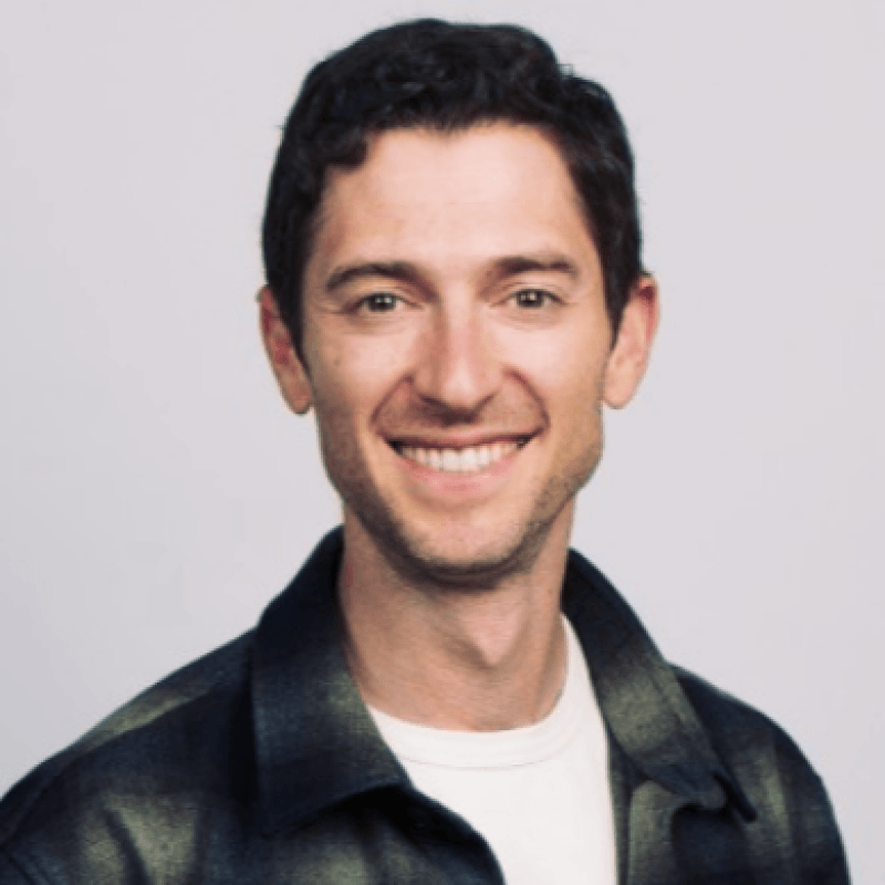 A man with short dark hair smiles at the camera, wearing a dark plaid shirt over a white t-shirt against a plain light background—Jake Saper exudes approachable confidence, embodying domain credibility in both style and presence.
