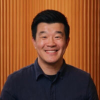 Smiling man with short black hair wearing a dark button-up shirt, standing in front of a background with vertical wooden slats, exuding confidence as he navigates digital onboarding processes to enhance Customer Success.