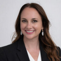 Woman with long brown hair wearing a black blazer, white blouse, and drop earrings, smiling in front of a plain light background—ready to lead your customer success onboarding with Hootsuite expertise.