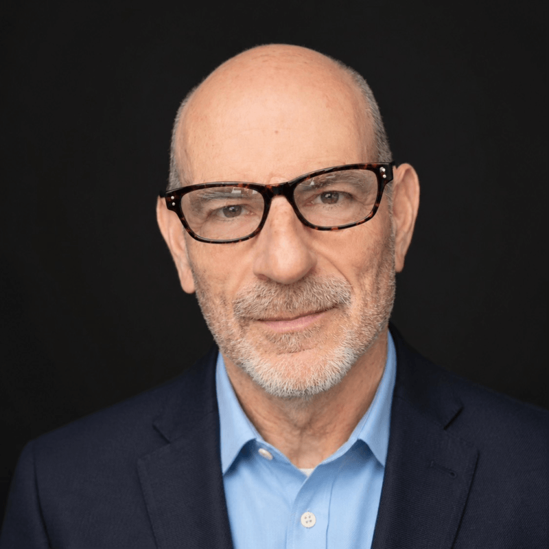 A bald man with glasses and a gray beard wearing a blue collared shirt and a dark blazer poses against a plain black background.