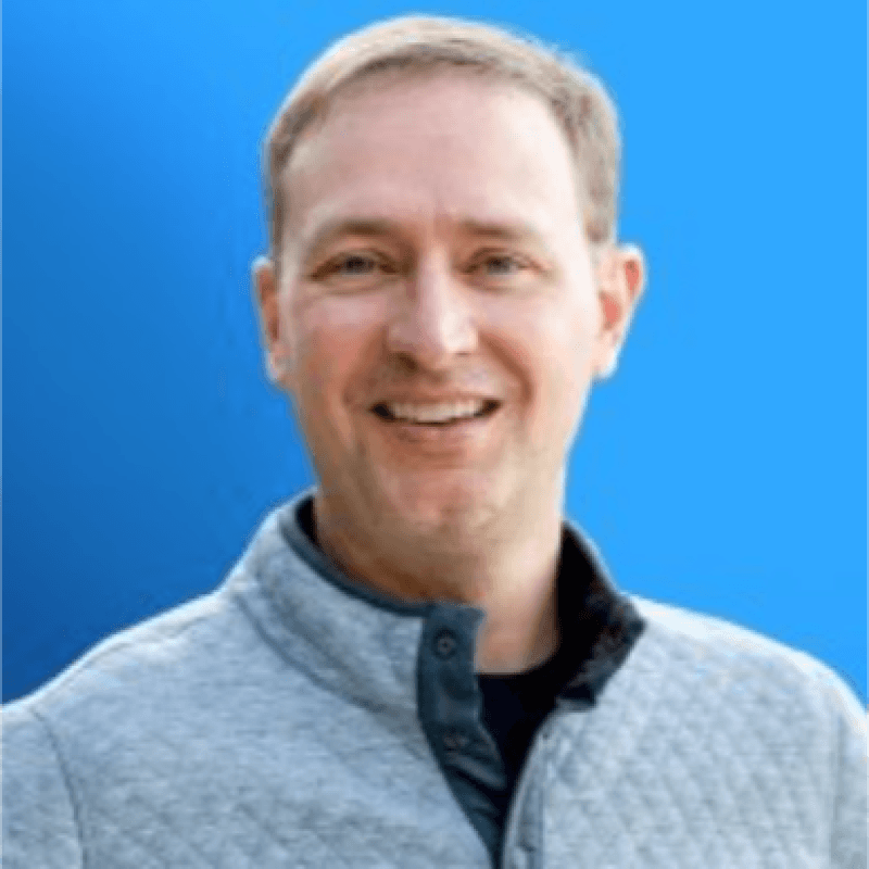 A man with short light brown hair smiles at the camera, wearing a light gray quilted jacket over a dark shirt, with a bright blue background—perfect for highlighting professionals like Andy Mowat who excel in VP roles or those looking to find VP jobs.