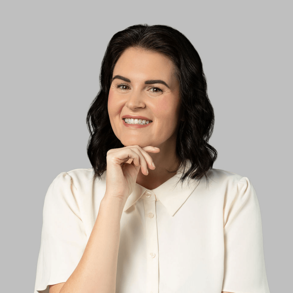 A woman with wavy dark hair, wearing a cream-colored blouse, smiles and rests her chin on her hand against a plain gray background.