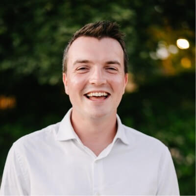 A man in a white shirt smiles broadly outdoors, with greenery and blurred lights in the background, embodying the positive energy often seen among Outcomes Architects and CSMs who use Notion to drive success.