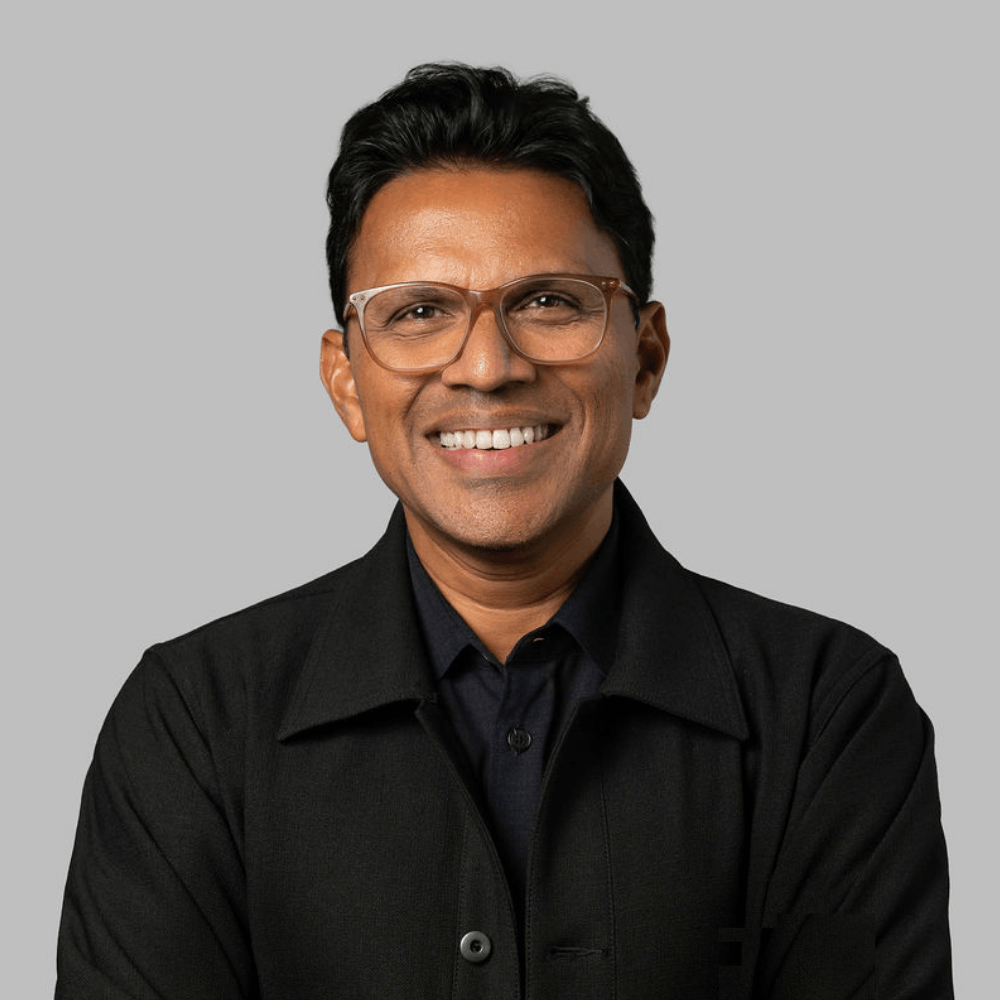 Smiling man with short dark hair, wearing glasses, a dark shirt, and a black jacket, standing against a plain light gray background.